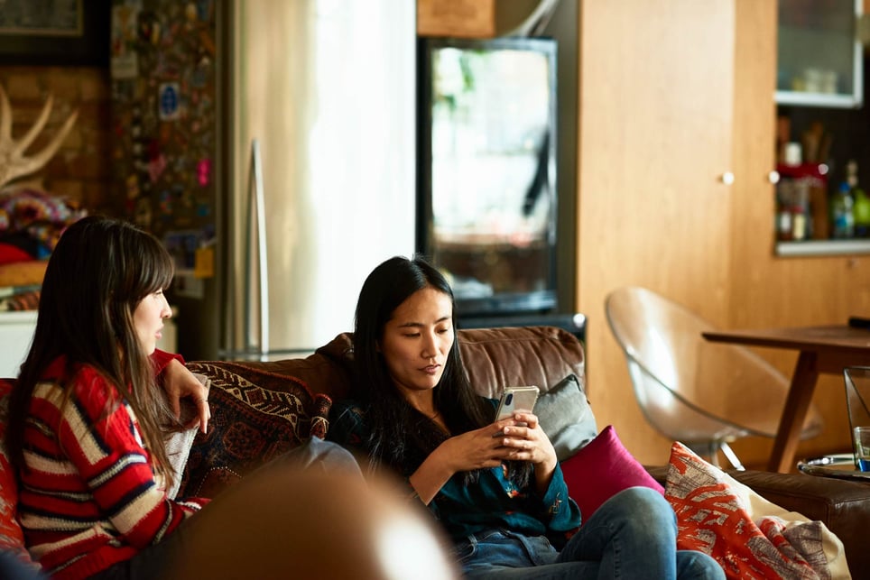 Chinese-woman-sitting-on-sofa-with-friend-using-cell-phone-listening-skills