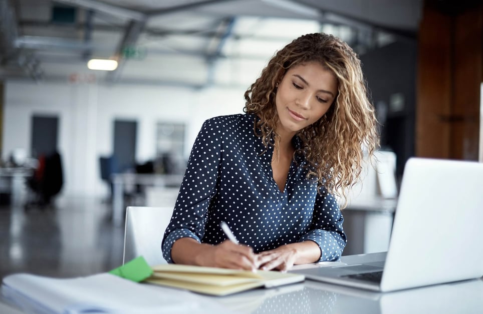 Young-woman-writing-in-a-notebook-in-front-of-her-laptop-how-to-write-a-professional-email