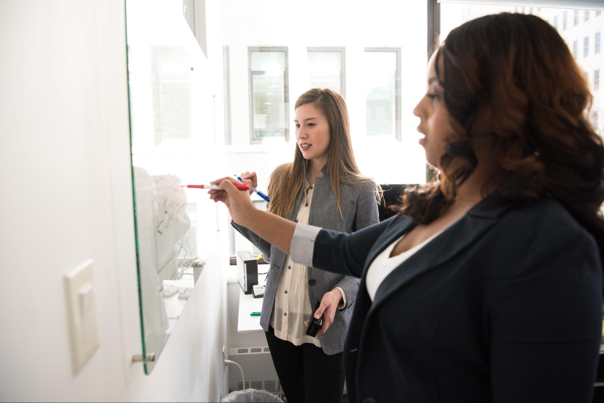 two-women-write-on-whiteboard-together-executive-development